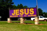 Em tradução, na placa está inscrita a frase "Jesus te dá boas-vindas à Hawkins".