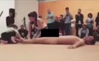 Criança de cerca de quatro anos (esquerda) é estimulada por mulher adulta (centro) a tocar corpo nu do homem que está deitado no tablado durante performance do '35º Panorama da Arte Brasileira – 2017'. (Imagem: Reprodução - Internet)