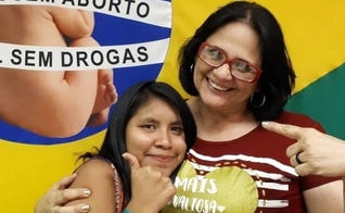 Damares Alves (direita) e Lulu (esquerda) abraçadas em frente à bandeira de uma campanha contra o aborto. (Foto: Instagram)