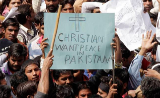 Homem segura em meio à multidão, cartaz que diz "Cristãos querem paz no Paquistão"