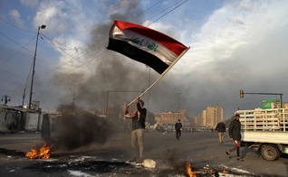 Manifestante acena a bandeira do Iraque enquanto fogo é ateado nas ruas próximas à Praça Tahrir, no Egito. (Foto: AP/Khalid Mohammed)