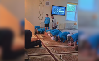 Jovem permanece em pé enquanto os demais participantes se ajoelham durante oração islâmica em mesquita. (Captura de tela/X/@megbasham)