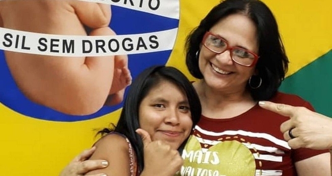 Damares Alves (direita) e Lulu (esquerda) abraçadas em frente à bandeira de uma campanha contra o aborto. (Foto: Instagram)