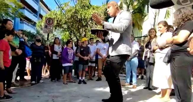 Com a Bíblia na mão, pastor fala aos participantes da reunião na porta da escola que sofreu tentativa de massacre. (Captura de tela/Tribunaonline)