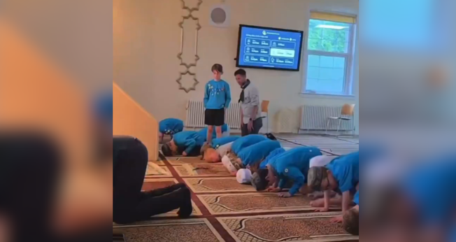 Jovem permanece em pé enquanto os demais participantes se ajoelham durante oração islâmica em mesquita. (Captura de tela/X/@megbasham)