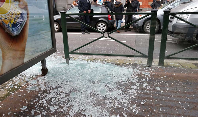 Policial olha para vidro quebrado no ponto de bonde, após a captura de suspeito de ataques em Bruxelas, enquanto a equipe de uma emissora local capta imagens. (Foto: Reuters)