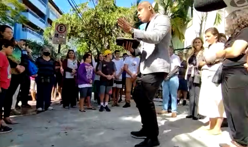 Com a Bíblia na mão, pastor fala aos participantes da reunião na porta da escola que sofreu tentativa de massacre. (Captura de tela/Tribunaonline)