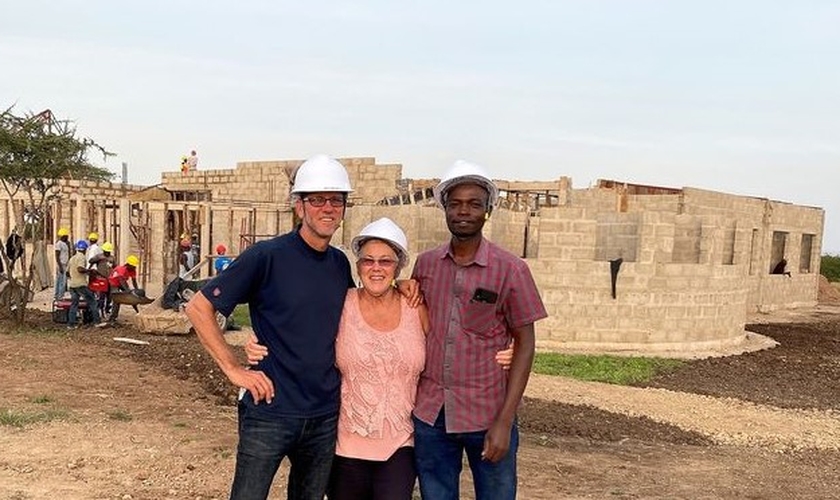 Darryl e Shirley Peters e auxiliar do casal, na Tanzânia; ao fundo, construção do centro de saúde. (Foto: Reprodução / CHVN Radio)